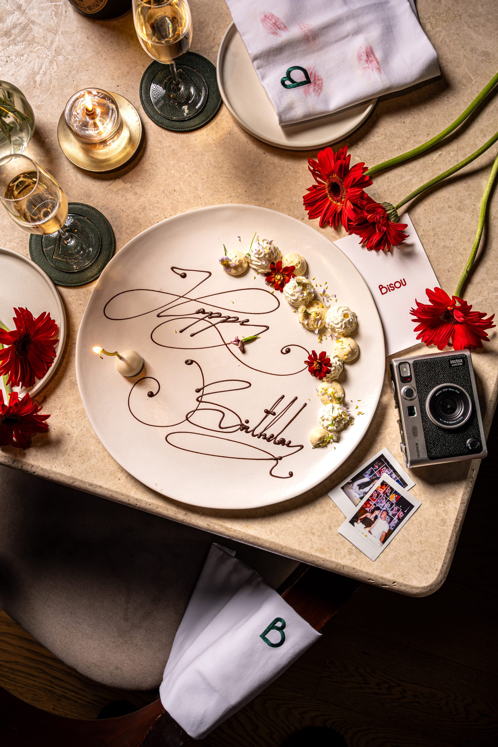 A Bisou birthday plate: Happy Birthday piped freehand in dark chocolate with quenelles, edible flowers and a lit candle, on a candlelit table