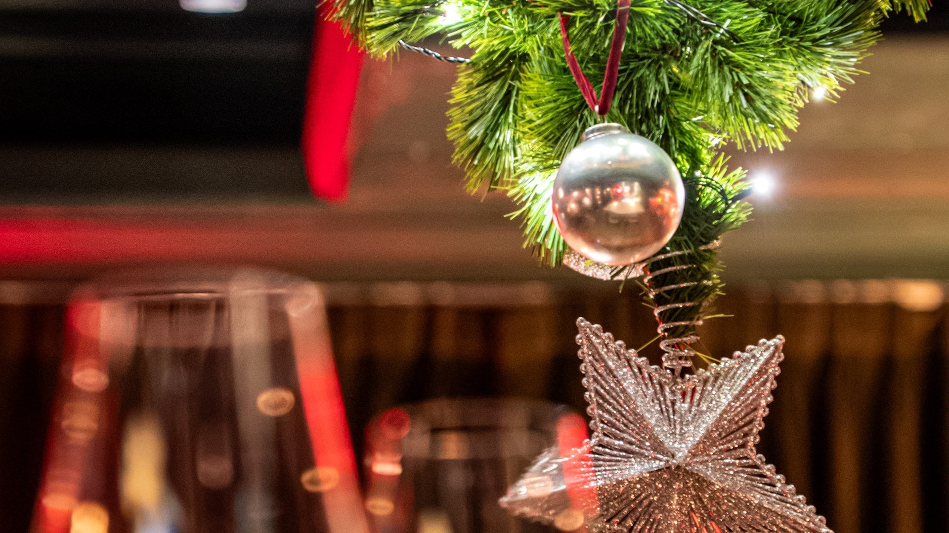 A silver bauble on a red ribbon and a glittering star hanging from the tip of a Christmas tree, with wine glasses blurred behind